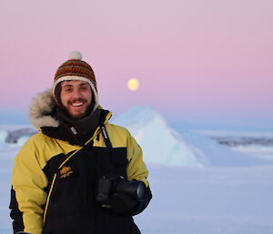 Person wearing a beanie and warm jacket standing in front of iceberg, with a full moon behind