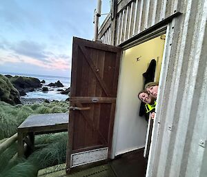 Two smiling women poke their heads around the front door of a corrugated metal field hut. Tussock grass and the ocean can be seen in the background.