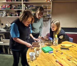 A woman shows two other women how to make custard slice. The women are standing and seated around a round dining table.