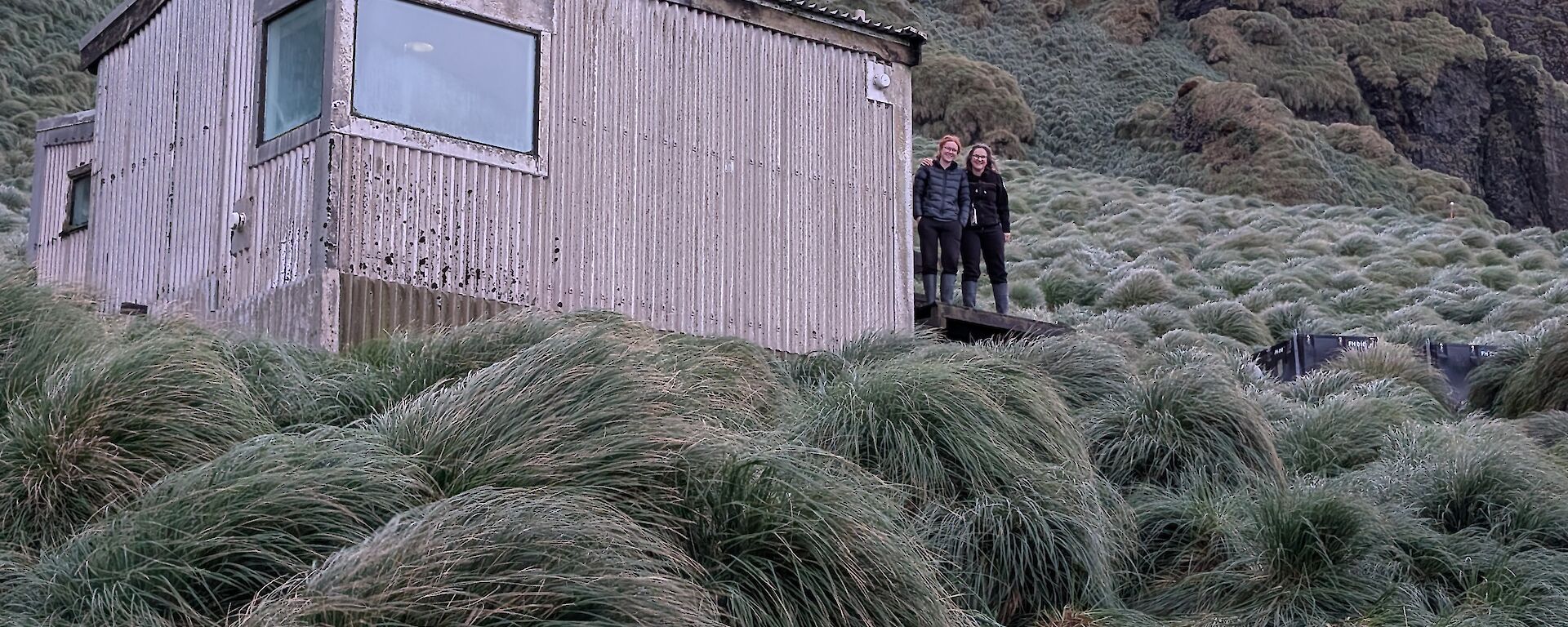 Two women stand by the side of a corrugated metal field hut nestled amongst the green tussocks.