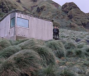 Two women stand by the side of a corrugated metal field hut nestled amongst the green tussocks.
