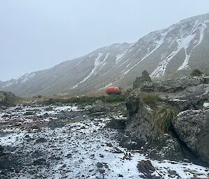 An orange spherical field hut sits amidst a snow-covered landscape. There is a steep range behind the hut.