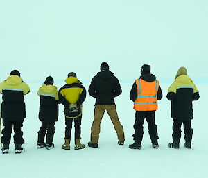6 expeditioners standing with their backs to the camera looking over the ice