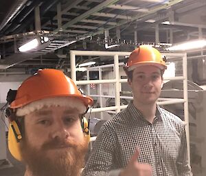 Two men standing in the winch room of RSV Nuyina, with orange hard hats on.