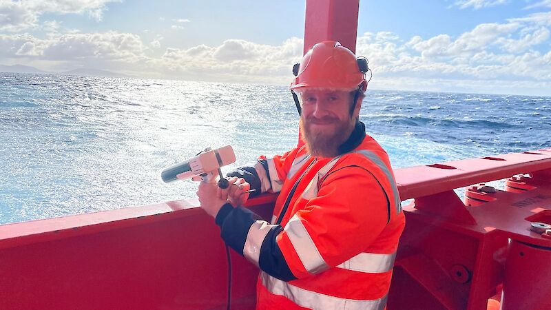 A man in a high visibility vest stands on the ship's deck with a small hand launcher for an ocean temperature probe, pointing out over the ocean.