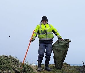 A smiling man in a yellow high-vis jacket holds an orange pole and a green canvas bag while standing amidst green tussock grass.