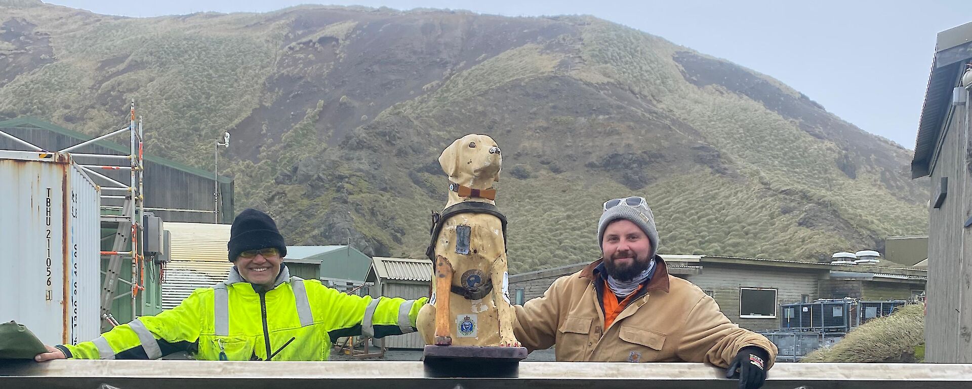 Stay, the Tasmanian collection Guide Dog sits atop the Maquarie Island gate with a smiling man standing on each side of her.