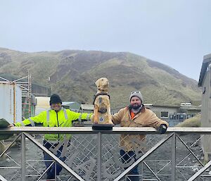 Stay, the Tasmanian collection Guide Dog sits atop the Maquarie Island gate with a smiling man standing on each side of her.