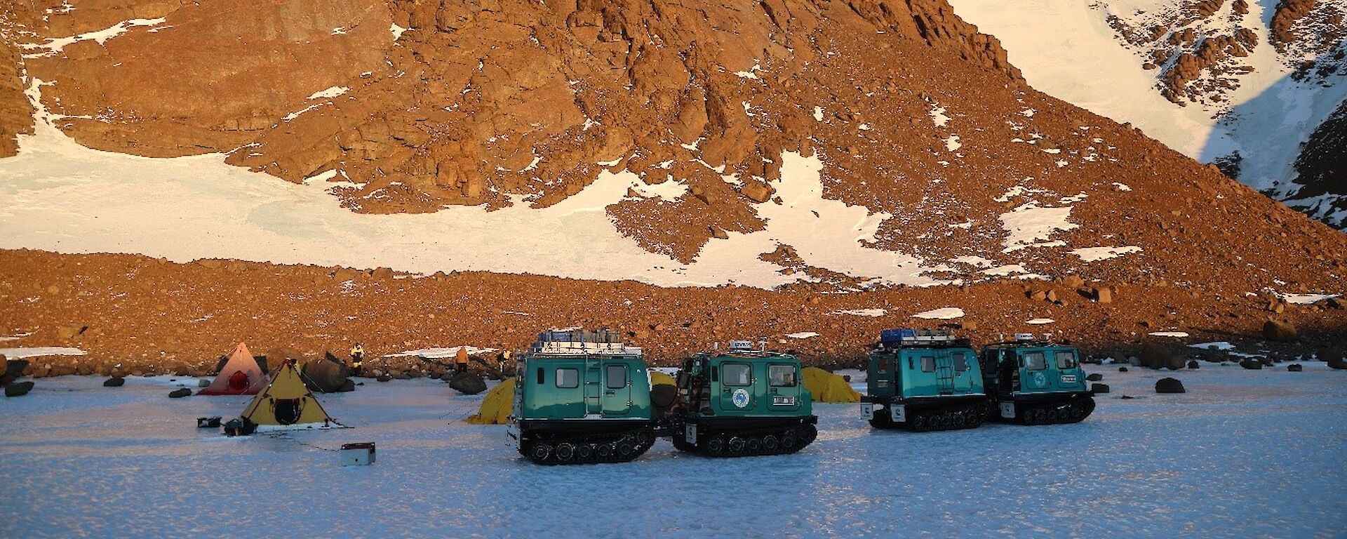 Two hagglund vehicles and polar pyramid tents on sea ice, with a rocky summit behind