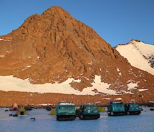 Two hagglund vehicles and polar pyramid tents on sea ice, with a rocky summit behind