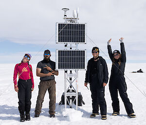 Four people standing next to a tall, steel, scaffolding-like structure carrying two solar panels and other scientific instruments