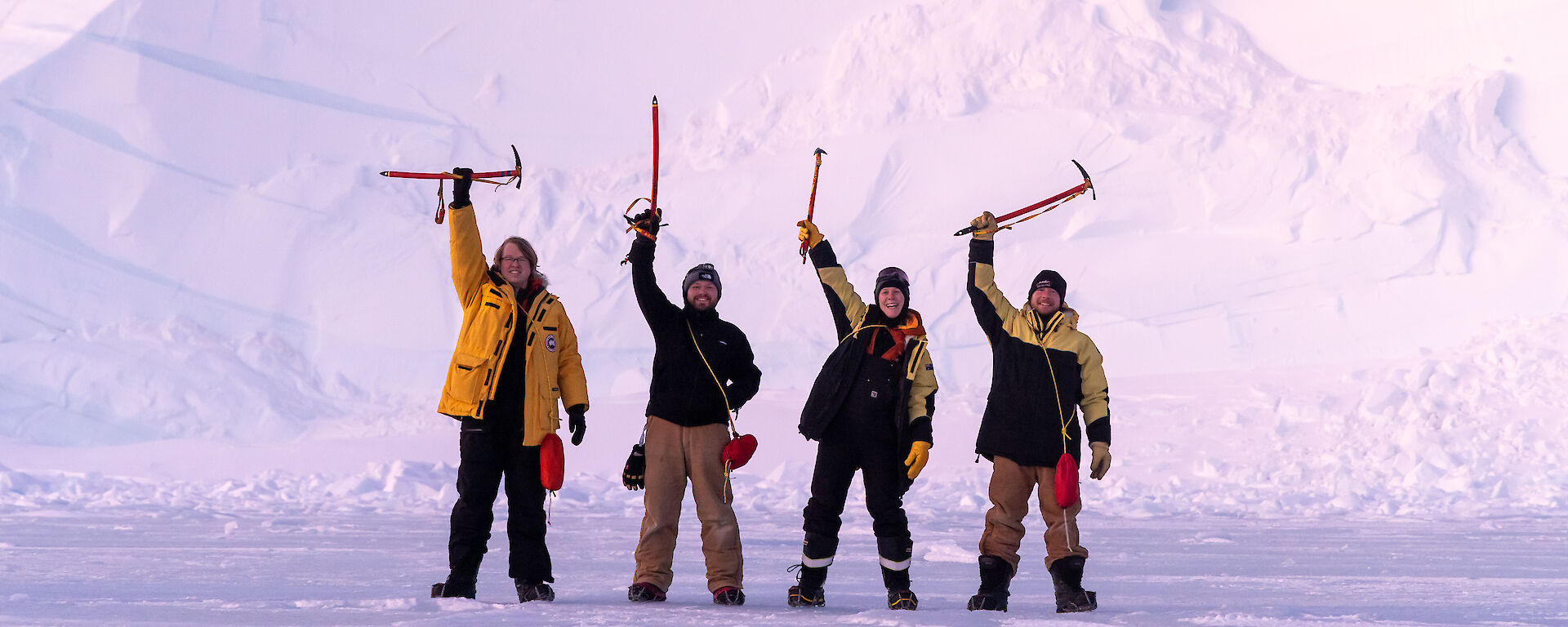 Four people standing in front of an ice cliff with their ice axes raised above their heads.
