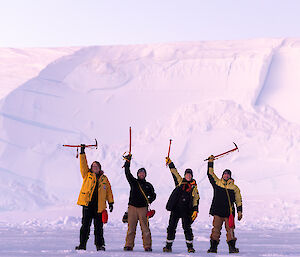 Four people standing in front of an ice cliff with their ice axes raised above their heads.