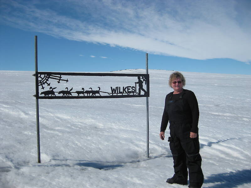 A woman dressed in black Antarctic clothing standing beside a sign for Wilkes, that includes cut-outs of huskies and a person behind a sledge.