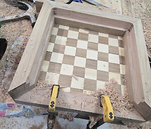 Two yellow clamps holding the dark and light wooden squares together that make up the chessboard.