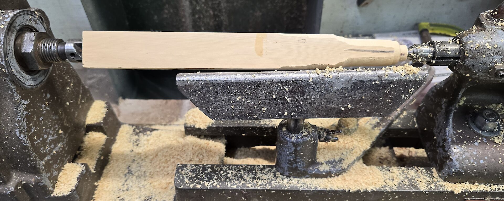 A length of square shaped Huon Pine sits in a lathe with some machining having commenced at the top end to create the beginnings of a chess piece.
