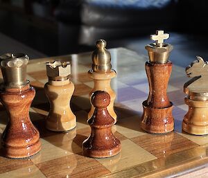 Six chess pieces - Queen, castle, pawn, bishop, king, horse - sit atop a wooden chess board. They have wooden bases made from wither Huon Pine or Blackwood and each is topped with a brass shape representing their role.