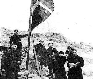Old photo of seven people standing around flag in Antarctica.