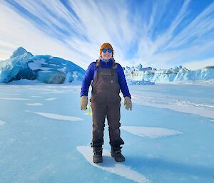 A woman in warm Antarctic clothing standing on sea ice, with jade icebergs rising above the ice behind her