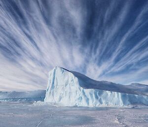An iceberg pushing up through sea ice, beneath a cloud-streaked blue sky