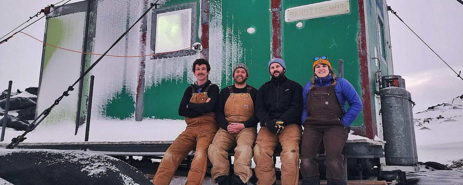 Four people sitting on the deck of a small, green field hut. The deck, hut and surroundings are covered in a thin layer of snow.