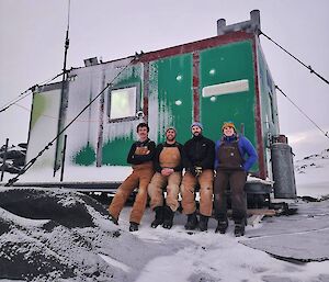 Four people sitting on the deck of a small, green field hut. The deck, hut and surroundings are covered in a thin layer of snow.