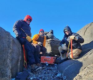 Three people sitting on rocks either side of gully, with an orange box between them