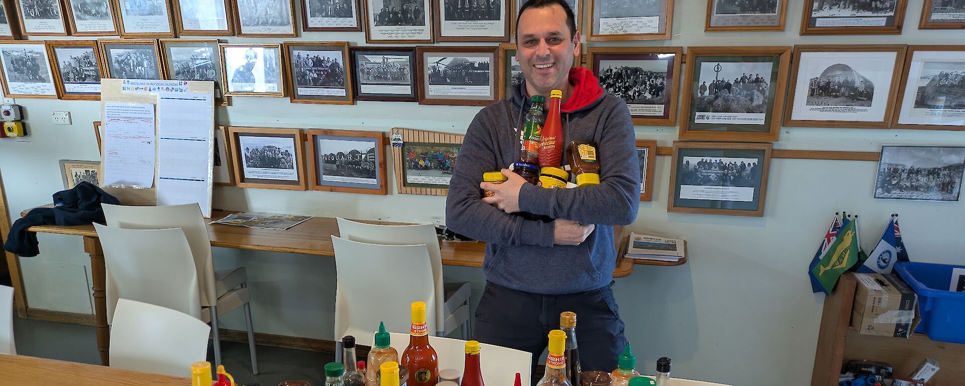 A smiling man holding bottles of various sauces, stands behind a table on which sit three trays filled with a broad range of sauces such as tomato, HP, BBQ, chilli etc