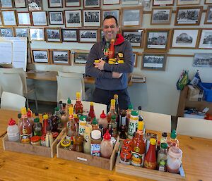 A smiling man holding bottles of various sauces, stands behind a table on which sit three trays filled with a broad range of sauces such as tomato, HP, BBQ, chilli etc
