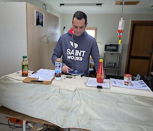 A man stands behind a work bench on which sit bottles of chilli sauce, HP sauce, BBQ sauce and mustard