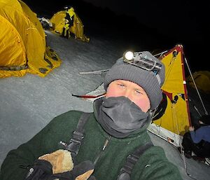 A man taking a selfie at night, in front  of three yellow tents pitched on sea ice