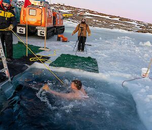 A man taking the plunge into a hole cut in the sea ice, while another person holds a rope attached around the swimmers chest