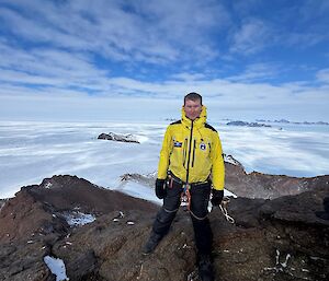 A man standing on top of a rocky summit, with sea ice and rocky peaks behind