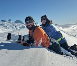 Two people lying on their stomachs in the snow. The man in front is holding a camera with a very long lens.