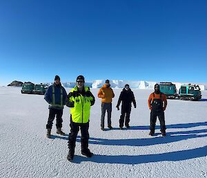 Five people standing on sea ice with two Hagglund vehicles in the distance behind them