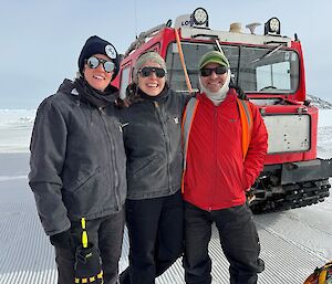 Three people stand on the ice in front of a haggland snow tractor