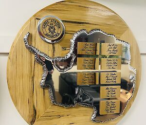 A round wooden plaque with a silver shape of Antarctica and a list of names of the 78th ANARE