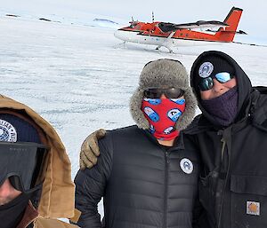 Three people, two wearing sunglasses and buffs over their noses, and one wearing ski googles, stand in front of a Twin Otter aircraft
