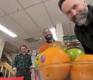 Oranges and apples in a glass bowl with three expeditioners in the background smiling at the fresh food