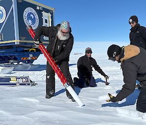 A man holds the barrel of a hand-held ice core drill at an angle to let a core of snow fall out the end. Three other people look on. One is holding part of the snow/ice core that has broken off.