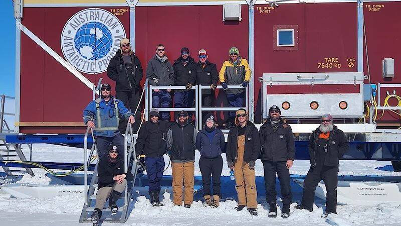 A group of 12 people standing in front of a large, red shipping container van on a sled, preparing to depart on a three-week traverse.