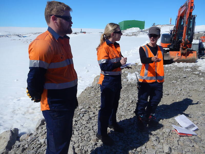 Two women and a man wearing orange high vis shirts, standing on a gravelly patch in some snow. A grader is in the background behind them.