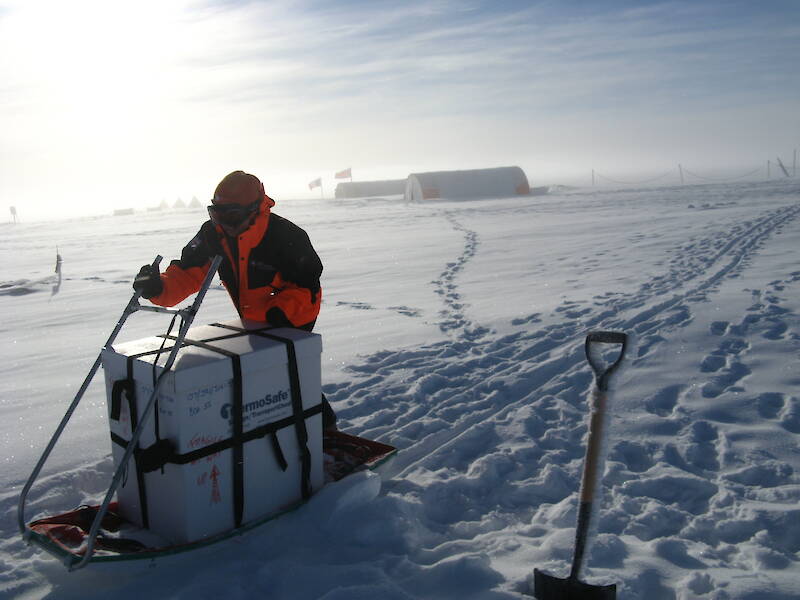 A woman pushing a box of ice cores on a sled across snow. Two large, cylindrical tents, used as shelters for ice core drilling, are visible in the distance behind her.