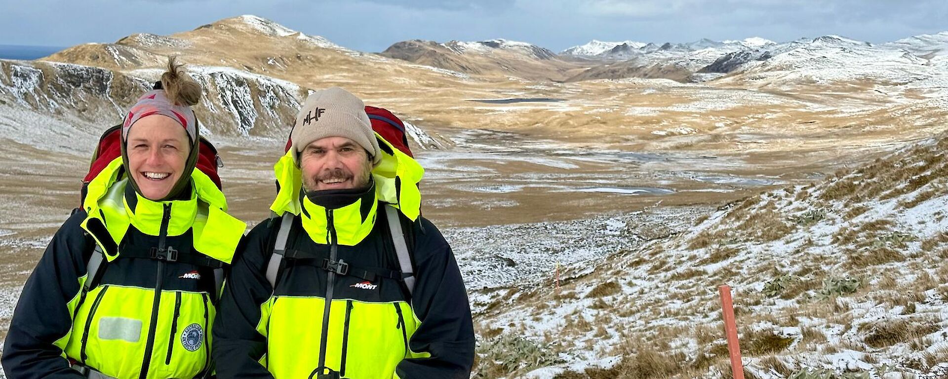 A smiling women and man wearing bright yellow jackets stand next to each other on a snow-covered landscape with many snow-capped peaks in the background.