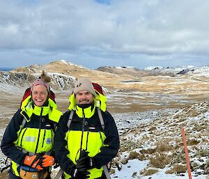 A smiling women and man wearing bright yellow jackets stand next to each other on a snow-covered landscape with many snow-capped peaks in the background.