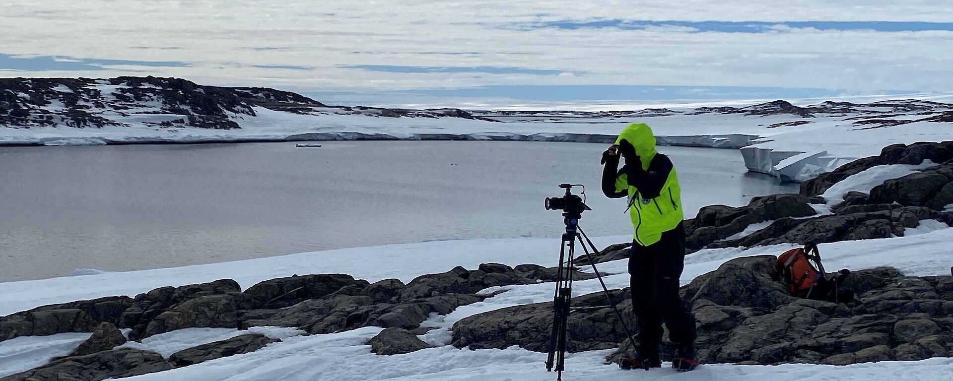 A person stands with a camera tripod on the ice amongst rocks with water and a headland in the background.