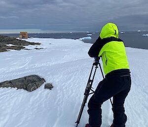 A person stands at a camera tripod looking out over ice and rocky terrain towards a structure with water and icebergs in the background