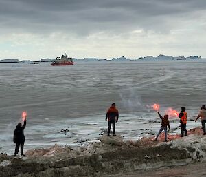 Five expeditioners on the beach at Davis waving farewell to a ship in the distance, Three of them have red flares in their hands.