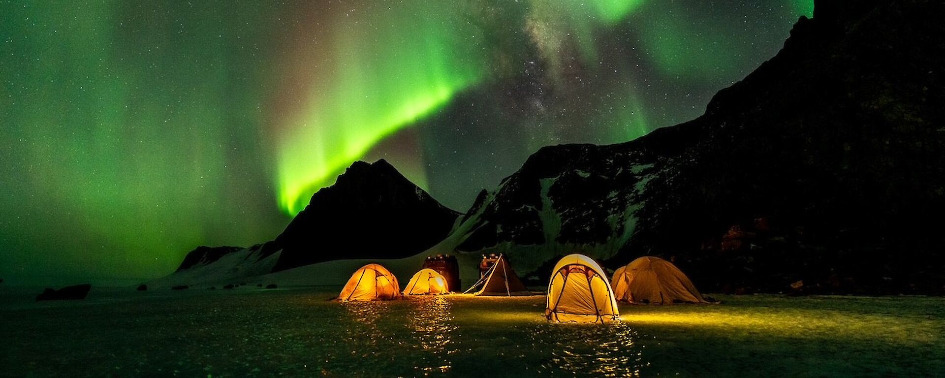 A brilliant green aurora in the sky above four yellow tents pitch on the ice. The tents are glowing from torchlight inside.