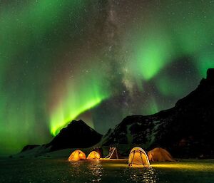A brilliant green aurora in the sky above four yellow tents pitch on the ice. The tents are glowing from torchlight inside.
